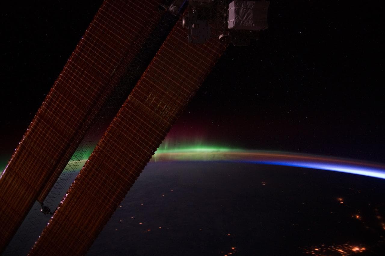 Earth's limb photographed from orbit at night: a green aurora arc curving above the dark horizon, a thin band of airglow along the edge of the atmosphere, and stars above it.